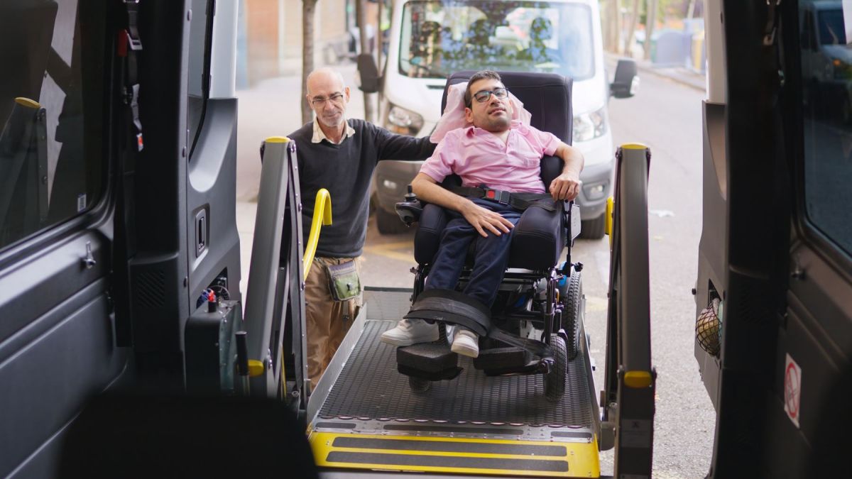 A driver helping a passenger in a power wheelchair board an accessible transit van by its lift
