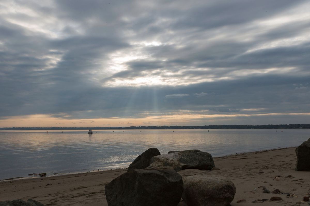 Sunlight breaking through clouds over a calm bay, with boulders on the beach in the foreground